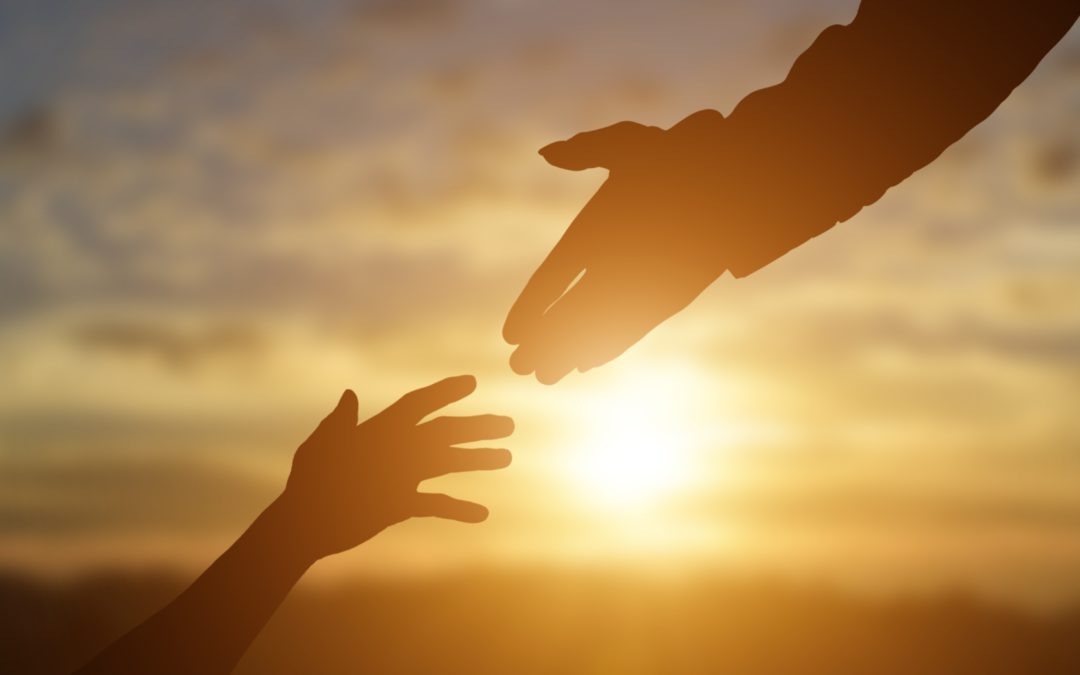 A silhouette of one hand reaching out to help another with a sunset in the background.